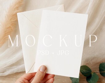 May include: A mockup featuring a blank white card and envelope, held by a hand. The card and envelope are off-white, with the word "MOCKUP" and "PSD + JPG" printed on the card. The background includes beige fabric, pampas grass, and eucalyptus leaves.