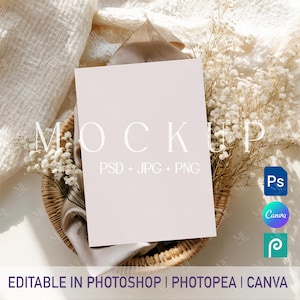 Könnte beinhalten: Eine leere, hellrosa Mockup-Karte mit dem Wort "MOCKUP" in Weiß, die in einem Weidenkorb mit Trockenblumen und Stoff liegt. Die Karte ist mit "PSD + JPG + PNG" und "EDITABLE IN PHOTOSHOP | PHOTOPEA | CANVA" beschriftet.