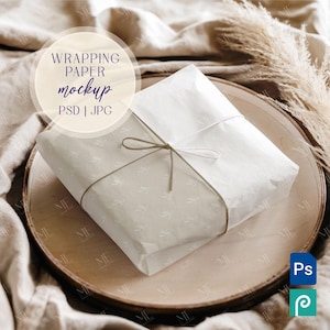Puede incluir: Un regalo blanco envuelto en papel de seda blanco con un simple lazo de cuerda, sentado en un plato de madera con un fondo de tela beige. El texto "WRAPPING PAPER mockup PSD | JPG" es visible en un círculo en la tela.