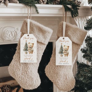 May include: Two beige Christmas stockings with a fluffy texture hang from a mantel. Each stocking has a tag that reads "Thank you" and features a teddy bear and Christmas tree illustration. The tags say "for showering our baby with love! David's Baby Shower."