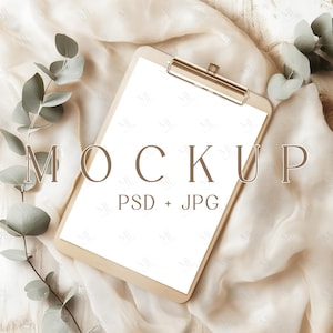 May include: A white clipboard with a gold clip, lying on a beige fabric background with green eucalyptus leaves. The text "MOCKUP PSD + JPG" is printed on the clipboard.