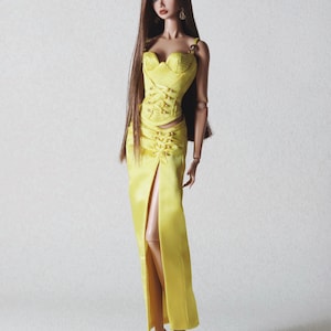 May include: A fashion doll with long brown hair wearing a bright yellow two-piece outfit consisting of a corset-style top with lace-up detailing and a matching floor-length skirt featuring a high front slit and lace-up accents.