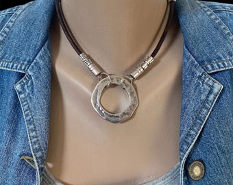 May include: A silver-toned necklace with a hammered, geometric pendant. The necklace features a dark brown cord and decorative silver accents. The pendant is a large, irregular circle. The necklace is displayed on a mannequin.