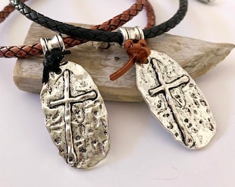 May include: Two silver-colored cross pendants with textured surfaces, each suspended from a braided leather cord. One cord is black, the other is brown. The pendants have a rustic, hammered appearance, and the crosses are centrally positioned.