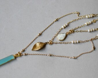 May include: A layered gold-tone necklace with multiple strands. The necklace features a rectangular, light blue pendant, a gold leaf charm, and a pear-shaped pendant. Small white pearls are interspersed along the strands. The necklace is displayed against a gray background.