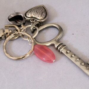May include: A silver-coloured keyring featuring a key, heart, and round charms, plus a pink oval bead. The key has a vintage design. The keyring has a lobster clasp and a textured key ring.