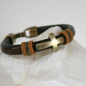 May include: A dark brown leather bracelet with a gold-colored cross and clasp. The bracelet features tan-colored accents and a decorative design. This accessory is suitable for everyday wear.