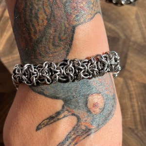May include: A silver-colored chainmail bracelet. The bracelet is made of interlocking metal rings, creating a textured, woven appearance. The bracelet is worn on a wrist with visible tattoos.