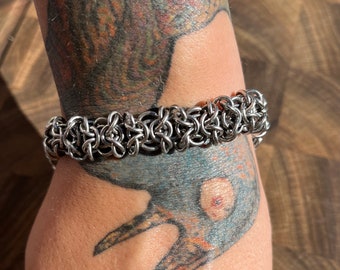 May include: A silver-colored chainmail bracelet. The bracelet is made of interlocking metal rings, creating a textured, woven appearance. The bracelet is worn on a wrist with visible tattoos.