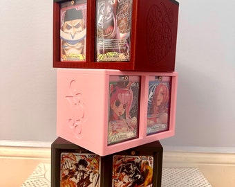 May include: A stack of three black, pink and red card display boxes with clear acrylic windows. Each box has two slots for displaying cards. The top box has a red heart design, the middle box has an anchor design, and the bottom box has a black and white design. The cards in the boxes feature characters from the anime One Piece, including Monkey D. Luffy, Sabo, Boa Hancock and Trafalgar D. Water Law.
