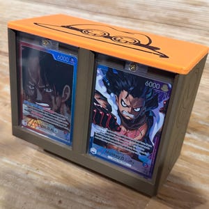 Puede incluir: Una caja de almacenamiento de tarjetas marrón y naranja con dos ranuras para tarjetas. La caja presenta ilustraciones de personajes de anime y el número 6000. La parte superior de la caja tiene un diseño decorativo.