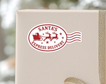 May include: A red and white oval sticker with the text "SANTA'S EXPRESS DELIVERY" and an illustration of Santa's sleigh and reindeer. The sticker is affixed to a beige gift box with a gold ribbon.