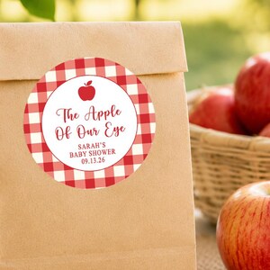 May include: A brown paper bag featuring a circular sticker with a red and white gingham pattern, a small red apple icon, and the text "The Apple Of Our Eye", "SARAH'S BABY SHOWER", and "09.13.26" printed in a dark red script and sans-serif font.