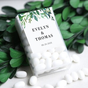 May include: A transparent rectangular box filled with white sweets. The box has a white label with the names "EVELYN & THOMAS" and the date "08.20.2025". The label is decorated with green watercolour leaves and berries. The sweets are scattered around the box.