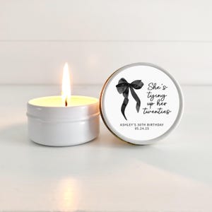 May include: A white candle in a tin with a black bow and the text "She's tying up her twenties" on the label. The label also includes the text "Ashley's 30th Birthday 05.24.25".