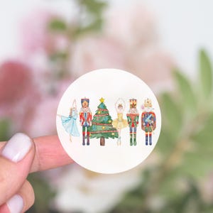May include: A round sticker featuring watercolor illustrations of a ballerina, a nutcracker soldier, a Christmas tree, another ballerina, a nutcracker soldier, and a mouse king. The sticker has a white background and a blurred background.