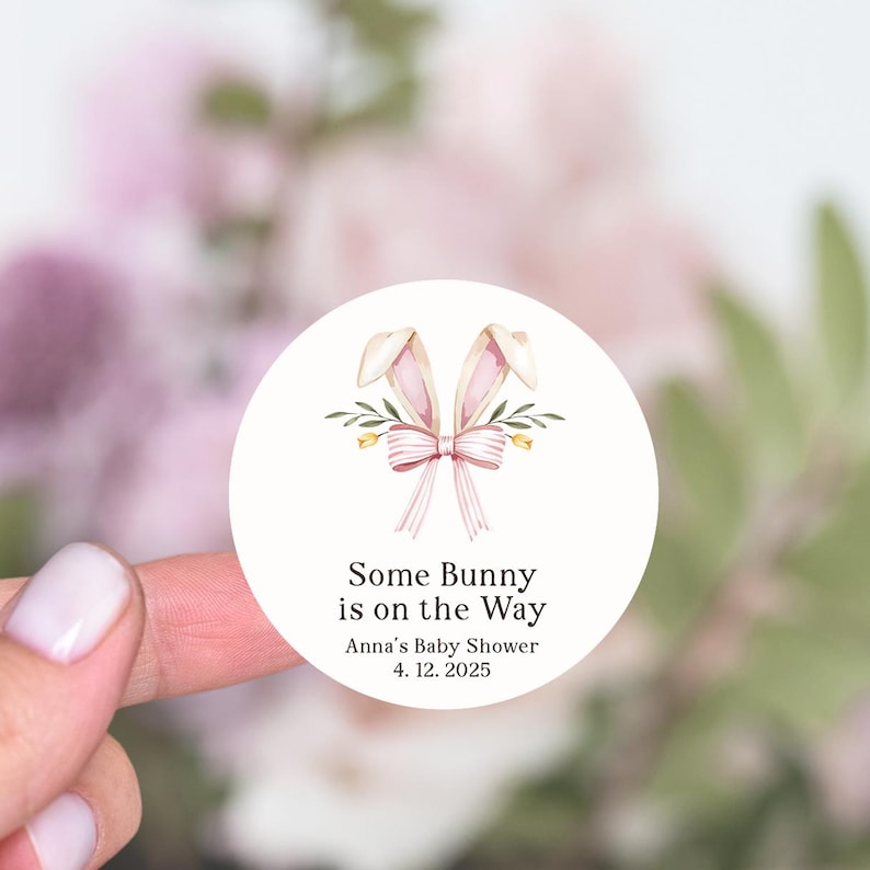May include: Round white sticker with a pink bunny rabbit ears and bow with green leaves and yellow flowers. The text reads "Some Bunny is on the Way" with "Anna's Baby Shower" and "4.12.2025" below.