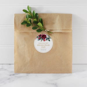 May include: A brown paper bag with a round sticker featuring a floral design and the text "Homemade by Emily Spiced Pear Jam Christmas 2021".