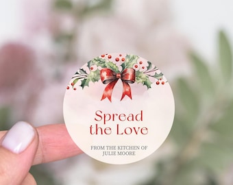 Puede incluir: Pegatina redonda con una ilustración de acuarela de un lazo rojo y hojas de acebo con bayas rojas. El texto "Spread the Love" está escrito en rojo debajo de la ilustración. El texto "From the kitchen of Julie Moore" está escrito en negro debajo del texto rojo.