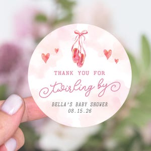 May include: A round sticker with a watercolor design. The sticker features pink ballet shoes, hearts, and the text "Thank you for twirling by" and "Bella's Baby Shower 08.15.26". The background is a soft pink watercolor wash.
