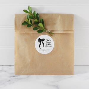 May include: A white round sticker with a black bow and the text "She's tying up her twenties" with the words "Ashley's 30th Birthday 05.24.25" below the bow.