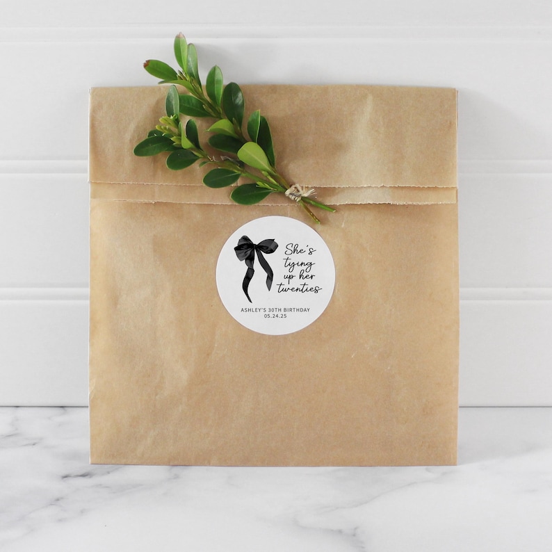 May include: A white round sticker with a black bow and the text "She's tying up her twenties" with the words "Ashley's 30th Birthday 05.24.25" below the bow.