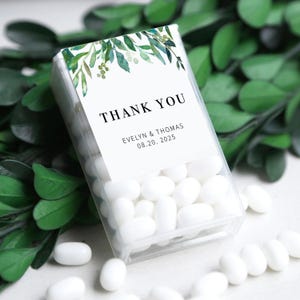 May include: A clear rectangular box filled with white sweets, labelled with "THANK YOU" and names "EVELYN & THOMAS" and the date "08.20.2025". The label features watercolor greenery. The box is surrounded by green leaves.