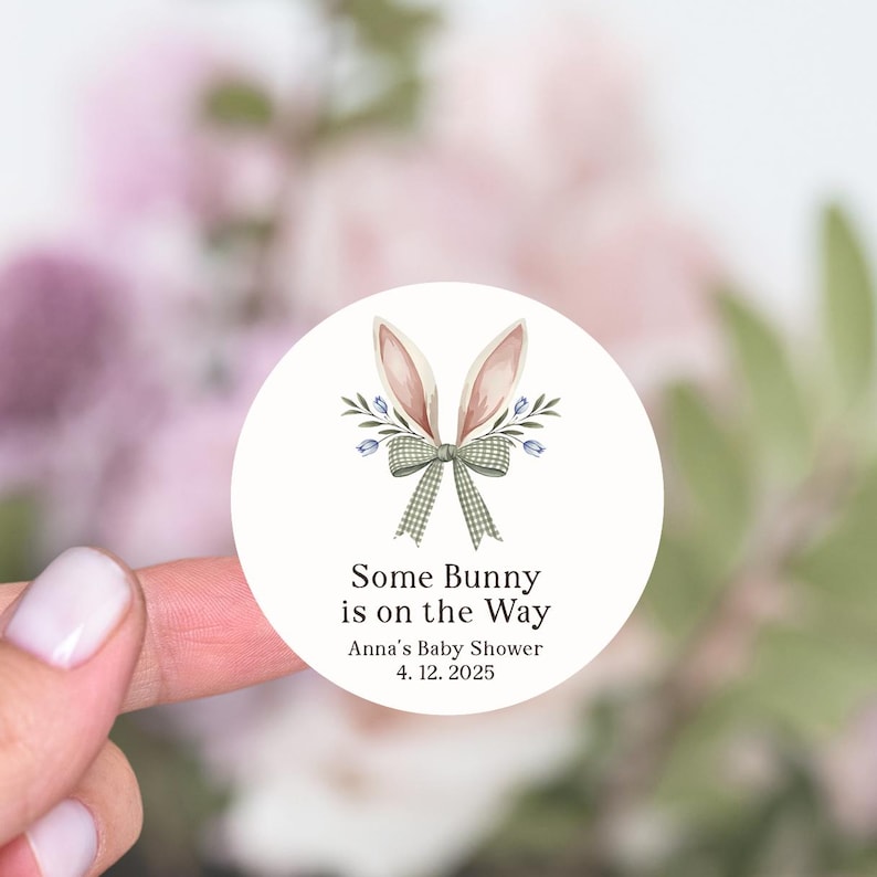 May include: White round sticker with a bunny rabbit illustration. The bunny has pink ears and a green and white checkered bow. The text reads "Some Bunny is on the Way" with "Anna's Baby Shower" and "4.12.2025" below.