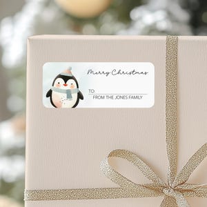May include: A white gift tag with a cartoon penguin wearing a blue scarf and a pink and white hat. The tag says "Merry Christmas" and "From the Jones Family".