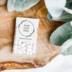 May include: A clear plastic box filled with white mints. The box has a white label with the text "YOUR LOGO HERE" inside a circular design and "WWW.WEBSITE.COM" below. The box is on a wooden surface with green foliage.
