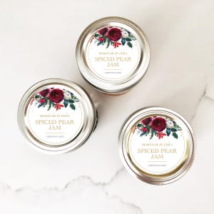 May include: Three glass jars with silver lids and white labels. The labels feature a red floral design and gold text that reads "HOMEMADE BY EMILY SPICED PEAR JAM CHRISTMAS 2021".