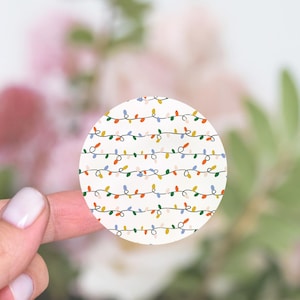 May include: Round sticker with a white background and a repeating pattern of colorful string lights. The lights are in shades of red, orange, yellow, green, blue, and pink.