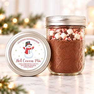 Op de afbeelding: Een glazen pot gevuld met bruin cacaopoeder, bedekt met witte marshmallows en stukjes pepermunt. Het zilveren deksel heeft een etiket met een sneeuwman en de tekst Olivia's Homemade Hot Cocoa Mix.