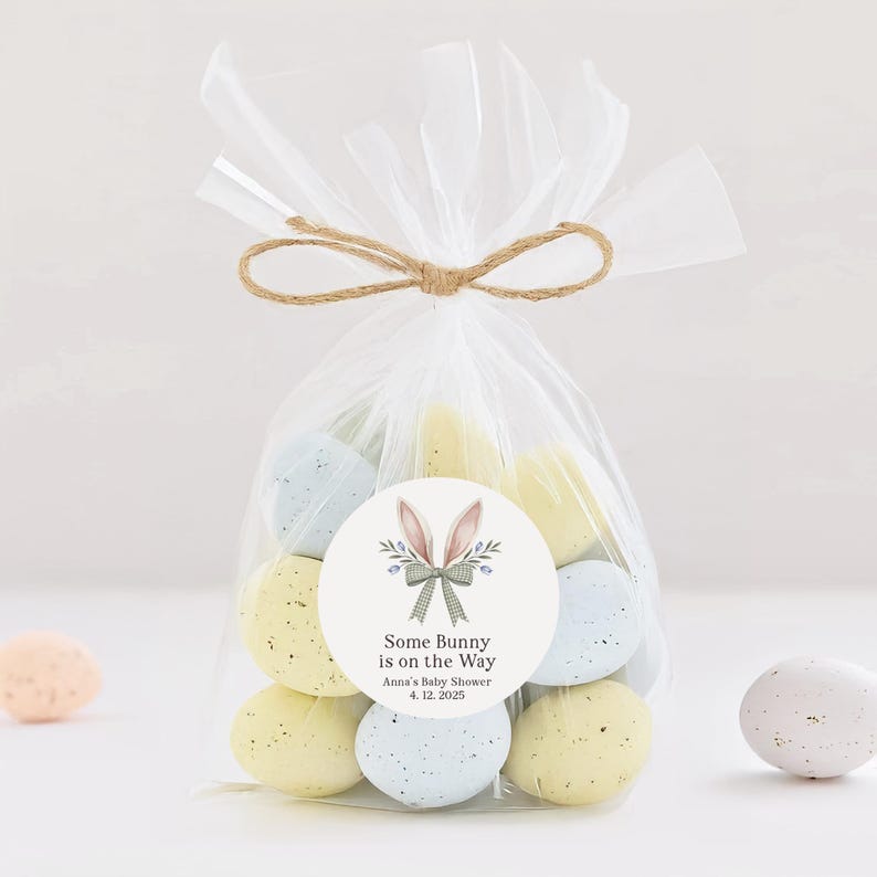 May include: A clear cellophane bag filled with pastel blue and yellow speckled Easter eggs tied with twine. A round sticker with a bunny rabbit illustration and the text "Some Bunny is on the Way" is attached to the bag. The text "Anna's Baby Shower 4.12.2025" is also on the sticker.