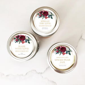 May include: Three glass jars with festive holiday labels. The labels feature a red and green floral design with gold accents. The jars contain homemade jams and jellies, including cranberry orange jam, spiced cranberry jelly, and spiced pear jam.
