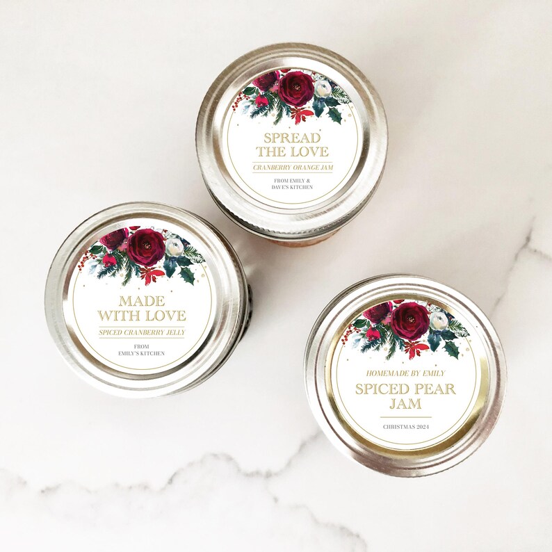 May include: Three glass jars with festive holiday labels. The labels feature a red and green floral design with gold accents. The jars contain homemade jams and jellies, including cranberry orange jam, spiced cranberry jelly, and spiced pear jam.