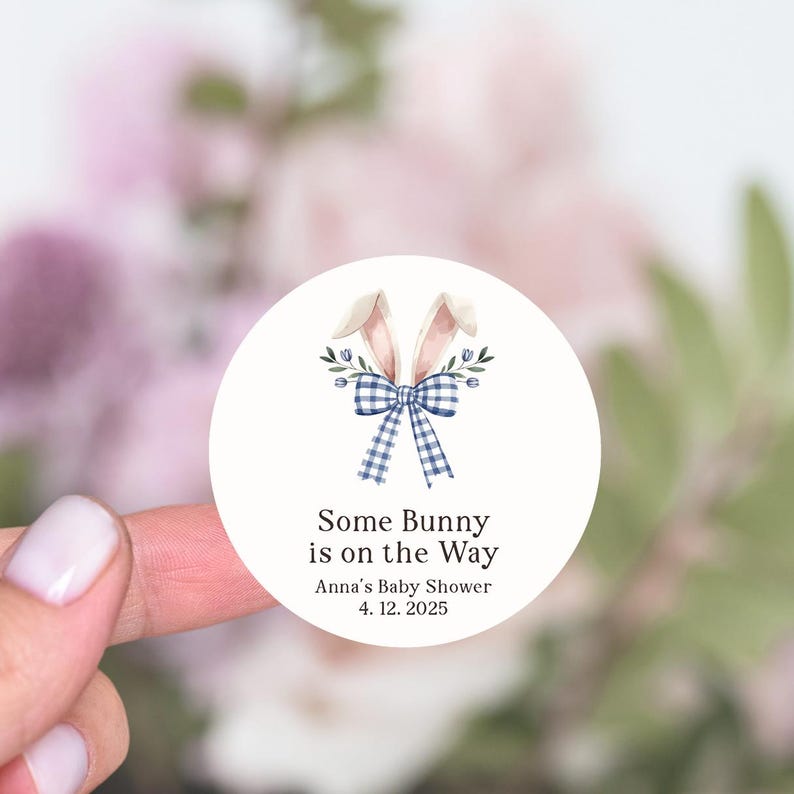 May include: Round white sticker with a blue and white gingham bow with bunny ears and green leaves. The text reads "Some Bunny is on the Way" with "Anna's Baby Shower" and "4.12.2025" below.