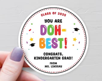 May include: A white circle sticker with red dotted border. The text reads "Class of 2025 You are DOH-BEST! Congrats, Kindergarten Grad! From Ms. Lindsay". The text "DOH-BEST!" is in colorful bubble letters.