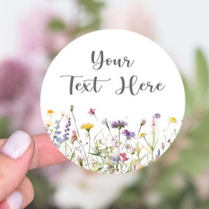 May include: A circular white sticker with the text "Your Text Here" in grey script. The bottom edge displays a watercolour illustration of wildflowers in shades of yellow, purple, and pink.