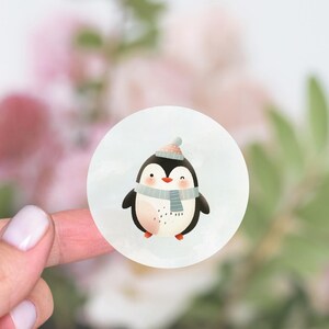 May include: A round sticker with a cute cartoon penguin wearing a blue and white scarf and a red and white hat. The penguin is on a white background.