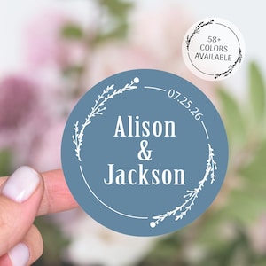May include: A round, dusty blue sticker with the names "Alison & Jackson" in white script, encircled by a white floral design. The date "07.25.26" is printed above the names. A smaller sticker reads "58+ COLORS AVAILABLE".