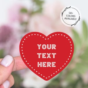 May include: A red heart-shaped sticker with white dotted edges and the text "YOUR TEXT HERE" in white. A circular sticker in the upper right corner says "58+ COLOURS AVAILABLE". The background is a soft, blurred floral arrangement.