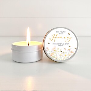 May include: A white candle in a tin container with a floral and bee design label that reads "She Found Her Honey, Emma's Bridal Shower 08.30.25"