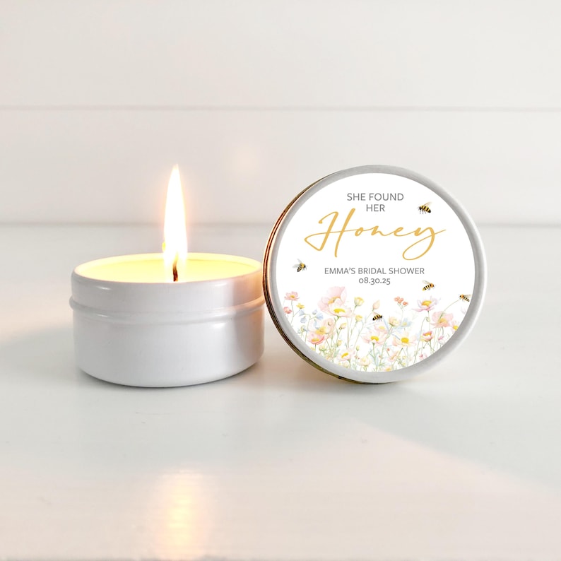 May include: A white candle in a tin container with a floral and bee design label that reads "She Found Her Honey, Emma's Bridal Shower 08.30.25"