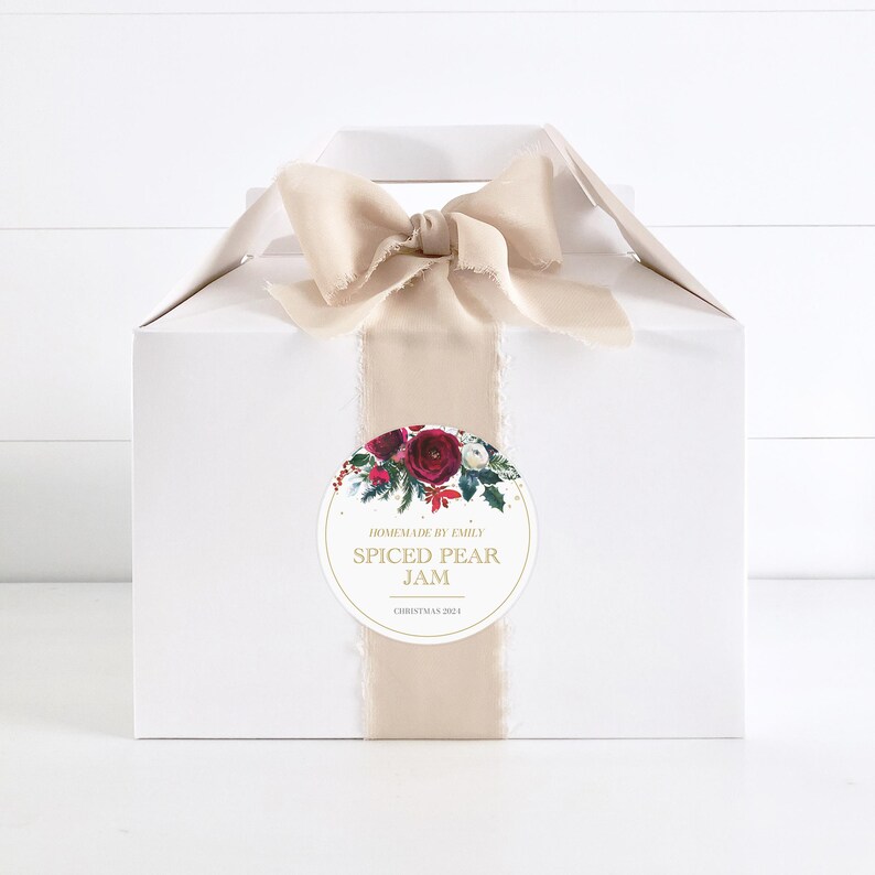 May include: A white gift box with a beige ribbon and a round sticker. The sticker has a red and white floral design and gold text that reads "Homemade by Emily Spiced Pear Jam Christmas 2021".