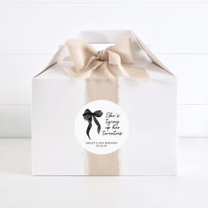 May include: White gift box with a beige ribbon and a round sticker that says "She's tying up her twenties" with a black bow illustration. The sticker also includes the text "Ashley's 30th Birthday 05.24.25".