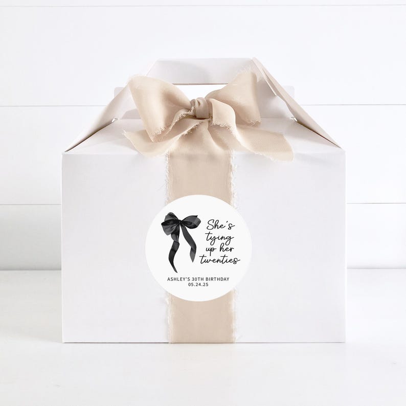 May include: White gift box with a beige ribbon and a round sticker that says "She's tying up her twenties" with a black bow illustration. The sticker also includes the text "Ashley's 30th Birthday 05.24.25".