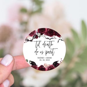 May include: Round wedding sticker with a floral design featuring dark red roses and bats. The text reads "Til death do us part" with the names Naomi and Adam and the date 10.18.2025.