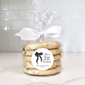 May include: A clear plastic bag filled with cookies, tied with a white ribbon. A round sticker with a black bow and the text "She's tying up her twenties" is attached to the bag. The sticker also includes the text "Ashley's 30th Birthday 05.24.25".