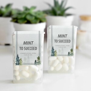 May include: Two clear plastic containers filled with white mints. Each container has a label that reads "MINT TO SUCCEED" with the text "CONGRATULATIONS, JAYDEN CLASS OF 2025" below. The labels feature watercolor pine trees.